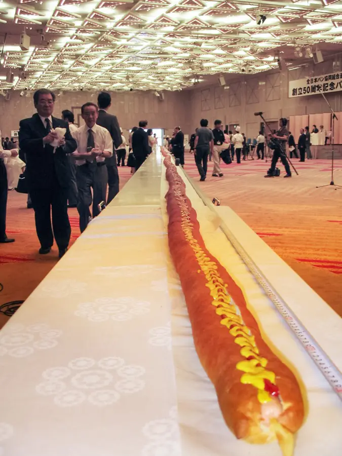 A hot dog sixty metres long runs the length of a banqueting hall on a white covered table, a line of mustard down it, with guests standing along both sides.