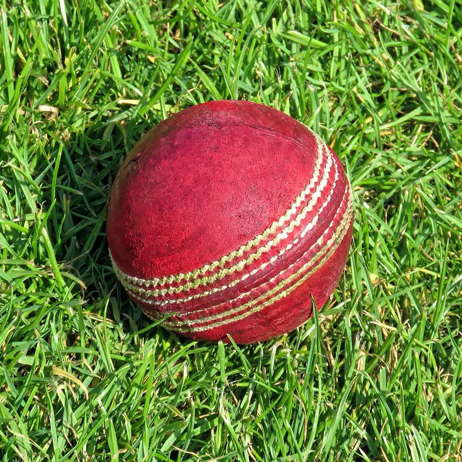 A used red cricket ball lying in green grass, seam and stitching worn. It is the first red on this page.